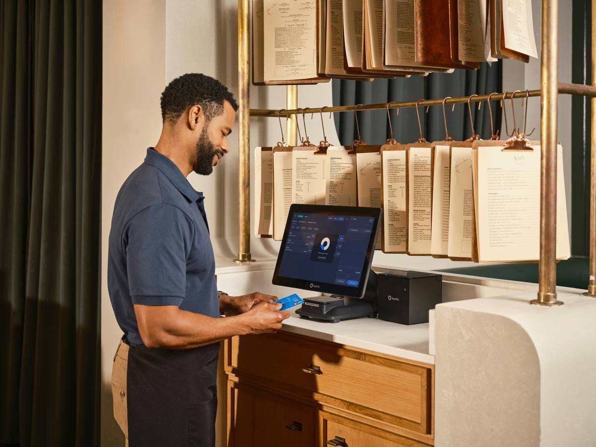 A server holds a credit card while processing a payment at a SpotOn countertop POS terminal at a sleek service station, with hanging menus on clipboards displayed behind him at an upscale casual dining restaurant. SpotOn's integrated payment processing gives restaurant staff the tools to handle transactions quickly and securely at every station. Learn more at spoton.com