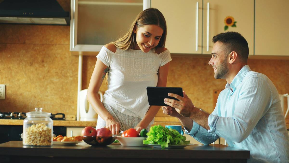 Attractive couple chatting in the kitchen early morning. Handsome man using tablet while his girlfriend cooking breakfast at home